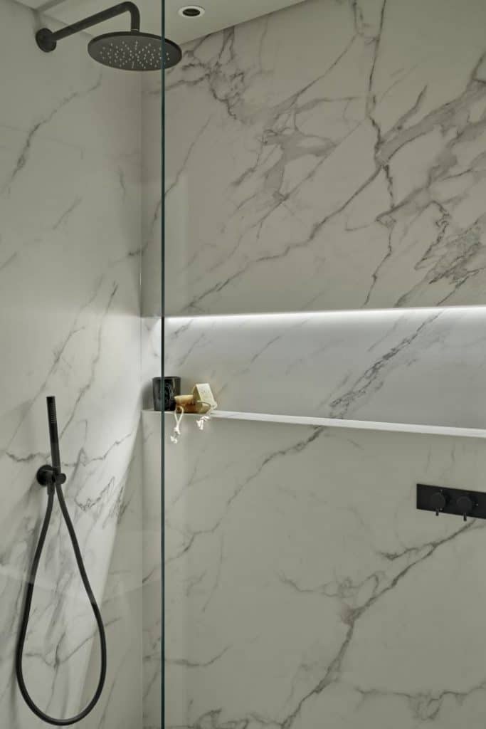 Guest bathroom with white marble effect porcelain tiles. the shower has a glass screen and black handwear, alongside a built in shelf accompanied with shower accessories.