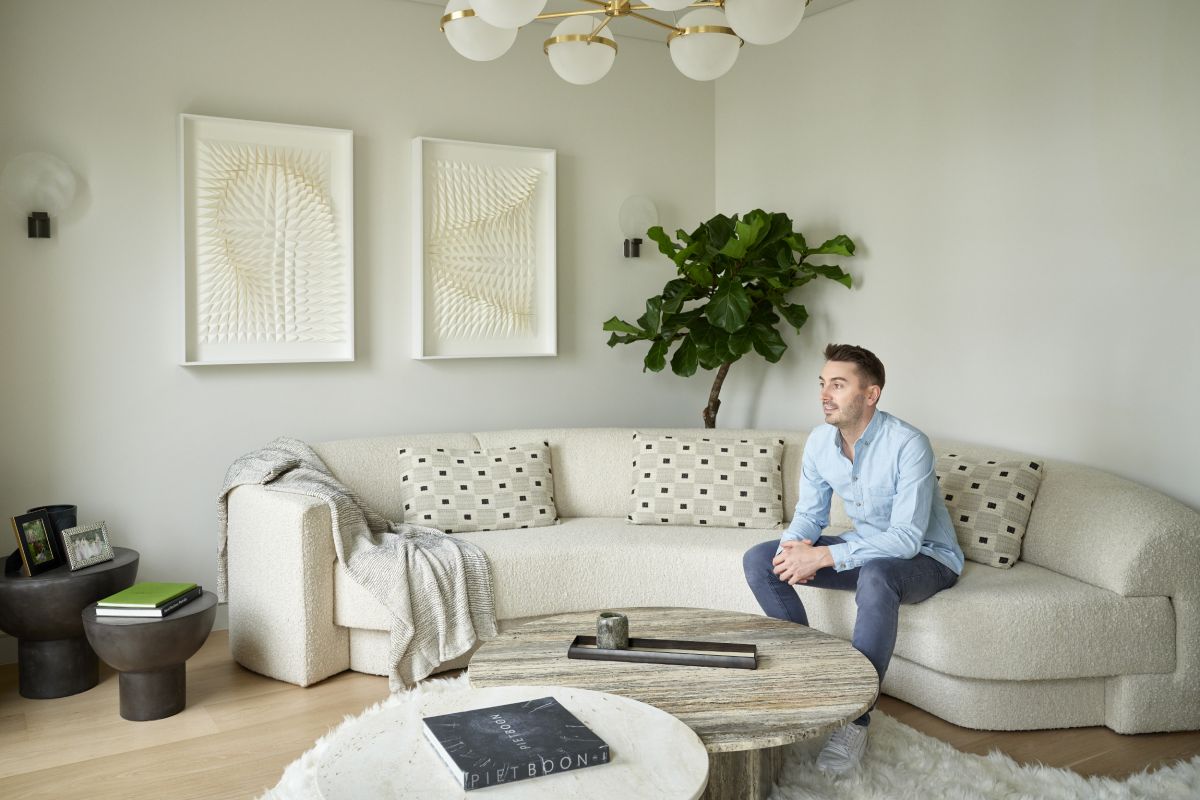 This luxurious living room features a corner space adorned with a curved white, textured sofa. Three matching white cushions and a white throw add comfort and elegance. Sitting on the sofa is renowned interior designer Brian Woulfe, responsible for the captivating design of this room. A fluffy curved rug lies in front of the sofa, hosting two marble tables in different natural tones. Adjacent to the sofa, two black side tables display books and framed photos. Adorning the wall are two white textured art pieces, while behind the sofa stands a tall, vibrant green plant, adding a touch of nature to the space.