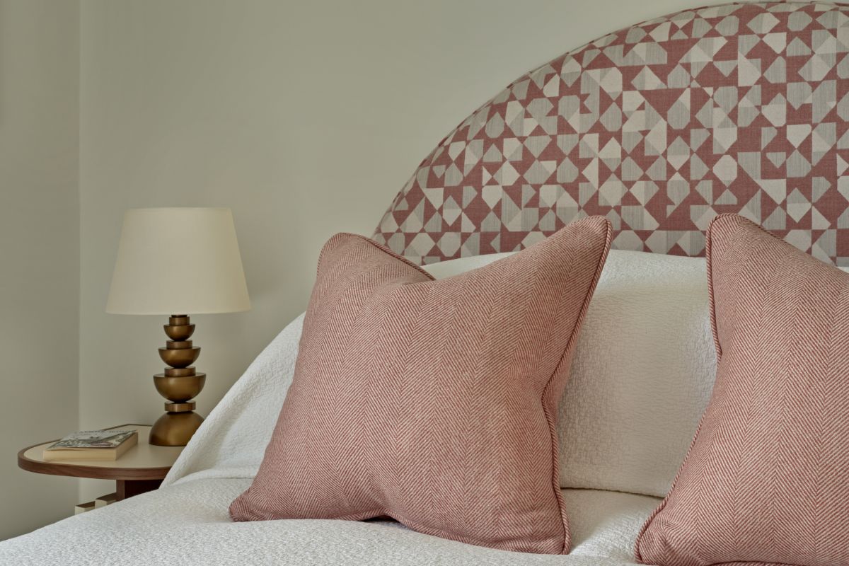 A luxury bedroom close-up image. The focal point is a magnificent bed with a curved pink and white geometric headboard, crafted from Holland and Sherry fabrics. Adorned with crisp white bedsheets and two small pink scatter cushions. Beside the bed, a circular wooden side table adds a touch of natural beauty, adorned with a book and an abstract lamp with a gold base.