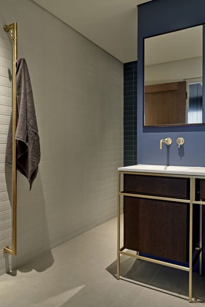 This is the epitome of a luxury bathroom. The grey stone tiled floor sets the stage for a serene atmosphere, complemented by the crisp white textured Marca Corona Regoli Porcelain wall tiles. A touch of elegance is added with the dark teal wall tiles on one side, while the sky blue painted wall adds a pop of bright colour. A centrepiece in the room, a large rectangular mirror hangs above a white sink with a brown wood base and gold metal pole structure, harmonising with the gold accented taps and towel rail. A dark grey towel hangs from the towel rail adding a homely, functional aspect to the setting.