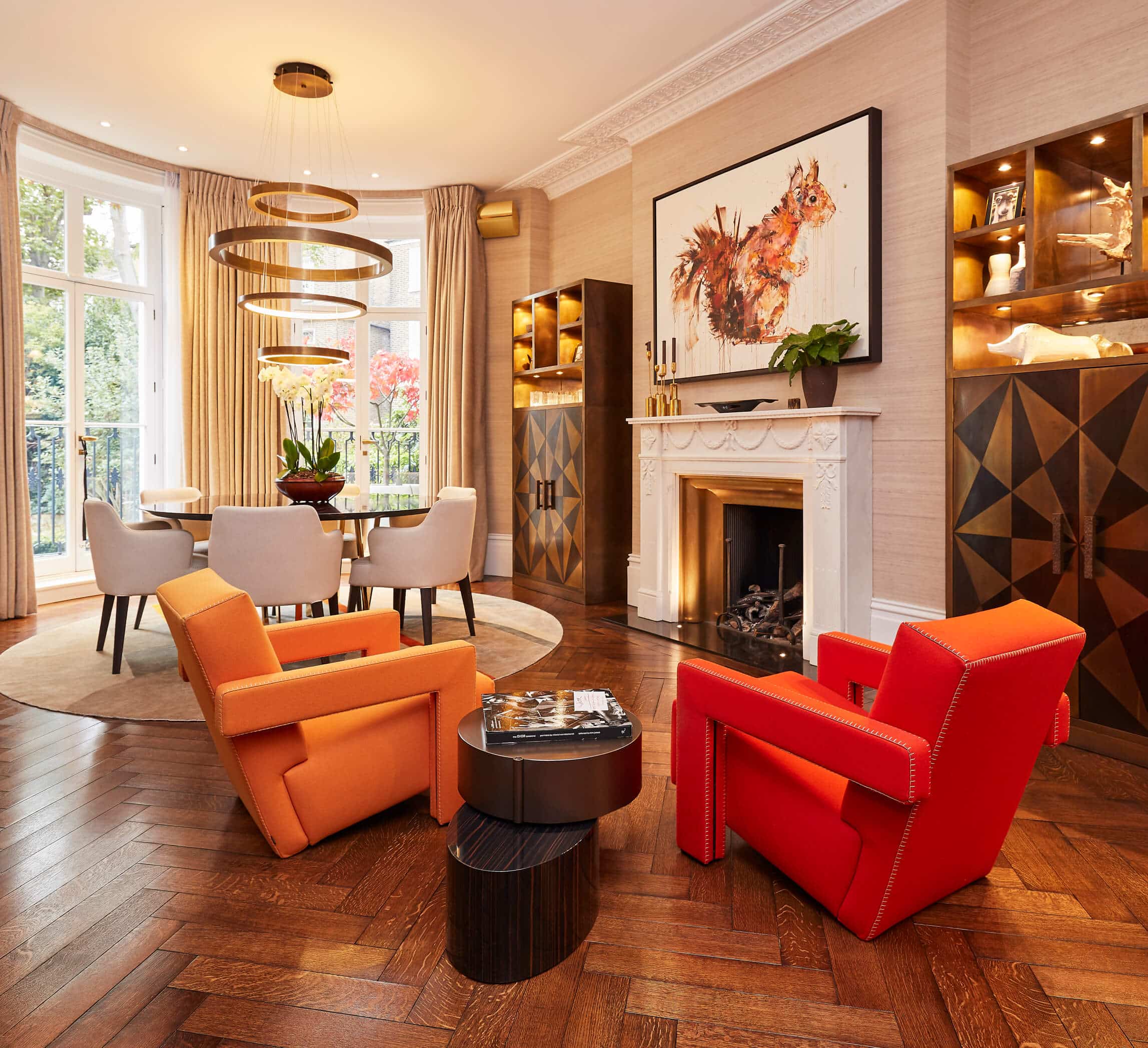 A luxury living room with a mid-century modern aesthetic, reminiscent of the stylish 80s design. The warm-toned herringbone wooden flooring adds a touch of warmth and sophistication to the space. Two Cassina Utrecht armchairs make a bold statement with one in bright red and the other in orange, creating a vibrant focal point. An abstract coffee table adorned with a book sits at the center. A white detailed fireplace with a gold interior exudes elegance, while a captivating piece of art hangs above it. Built-in cupboards on either side of the fireplace showcase geometric patterning, harmonizing with the gold and wood tones. A Sahara Noir circular dining table rests on a luxurious bespoke circular silk rug, surrounded by six white dining chairs with black legs, offering a stylish setting for entertaining guests. The living room is graced by two floor-length windows, adorned with curtains, which allow natural light to bathe the room, completing this luxurious space.