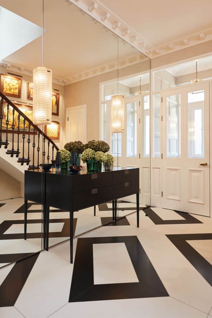 An exquisite hallway next to the front door. The venetian plaster walls exude elegance, while the black and white diamond-shaped tiles on the floor add a touch of charm. One wall is adorned with large panel mirrors, reflecting the beauty of the space. In front of the mirrors, a sleek console with pull out drawers showcases tasteful decor, including a collection of vibrant flowers. Above the console, two pendant lights glow. The front door, you'll be greeted by a grand, tall white door adorned with tall windows, inviting in natural light .