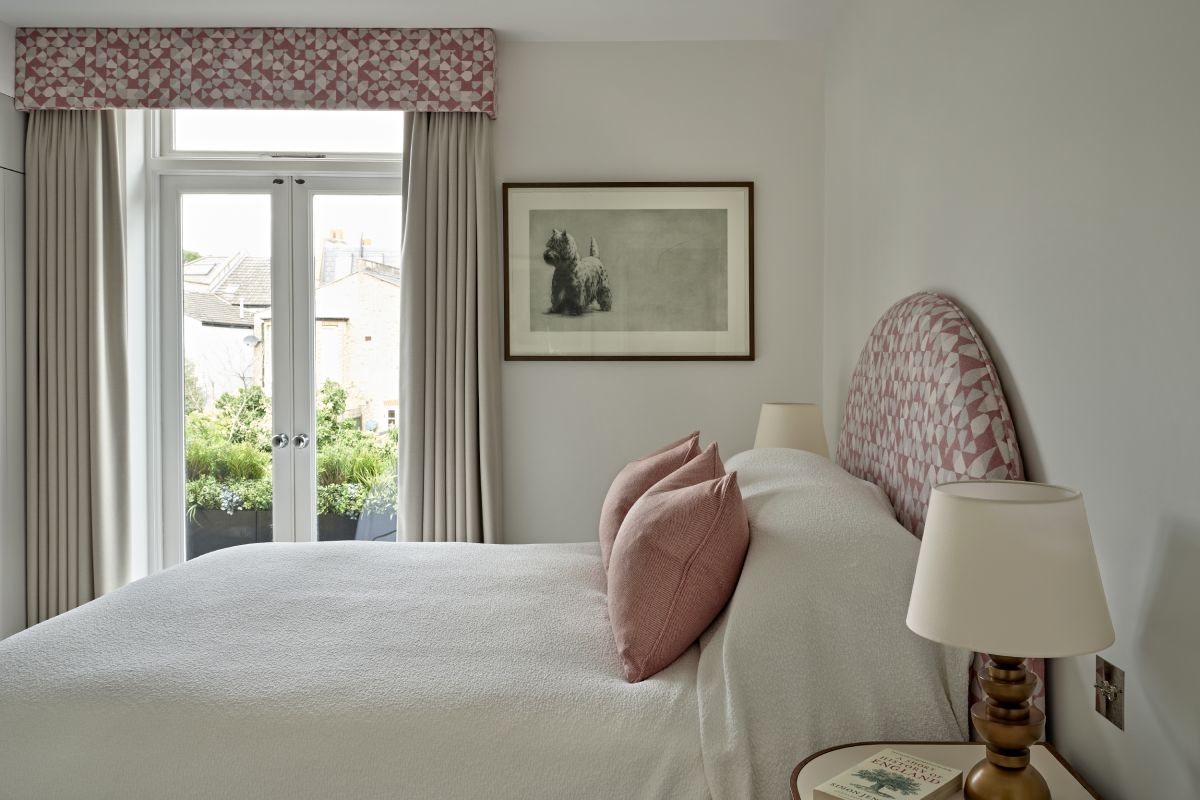 A luxury bedroom. The focal point is a magnificent bed with a curved pink and white geometric headboard, crafted from Holland and Sherry fabrics. Adorned with crisp white bedsheets and two small pink scatter cushions. Beside the bed, a circular wooden side table on either side of the bed adds a touch of natural beauty, adorned with a book and an abstract lamp with a gold base. Behind the bed a painting of a dog is framed on the wall next to large glass french doors that open to the beautiful green outside, the doors are adorned with cream curtains and matching fabric to the headboard covering the top of the curtains.