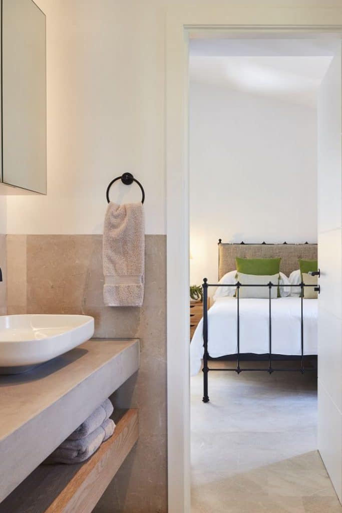 A guest bathroom finished with natural stone and wood and a micro cement vanity top leads into a bedroom with cast iron bed frame.