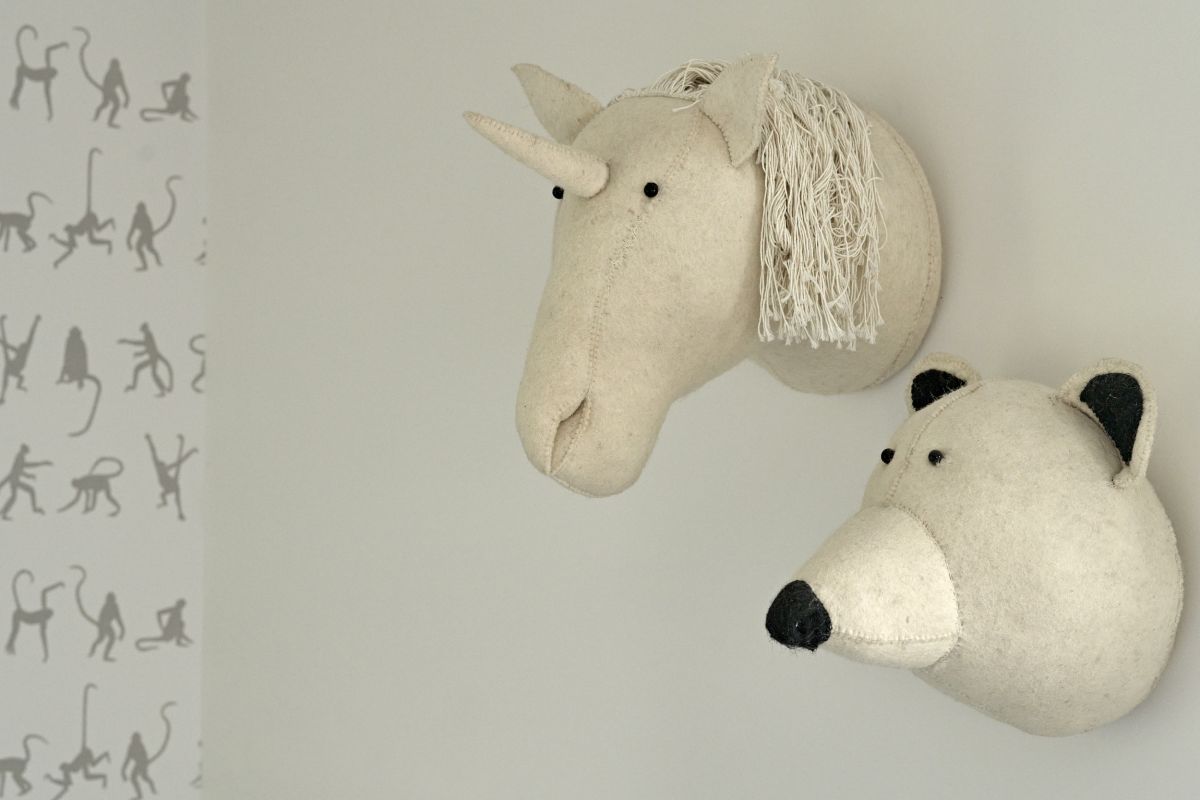 Close up image of whimsical stuffed animals heads hanging on children's bedroom wall, a fun cream unicorn and a matching bear head. in the background on another wall a monkey wallpaper, matching the animal theme.