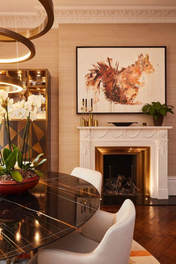 This luxury living/dining room exudes a timeless and sophisticated charm with its mid-century modern design elements. The dark wood herringbone flooring adds depth and richness to the space, while the warm-toned fishbone wooden flooring enhances its elegance. An abstract coffee table adorned with books becomes a focal point, complementing the overall aesthetic. The white detail fireplace with a gold interior adds a touch of luxury, with a captivating piece of art hanging above it. Built-in cupboards on either side of the fireplace feature geometric patterning, matching the gold and wood tones throughout the room. A black marble circular dining table placed on a cream rug becomes a statement piece, surrounded by white dining chairs with black legs. Orchids adorn the table, adding a touch of natural beauty. An ornamental gold ring light fixture suspended above the dining table completes the luxurious ambiance of this stunning living/dining room.