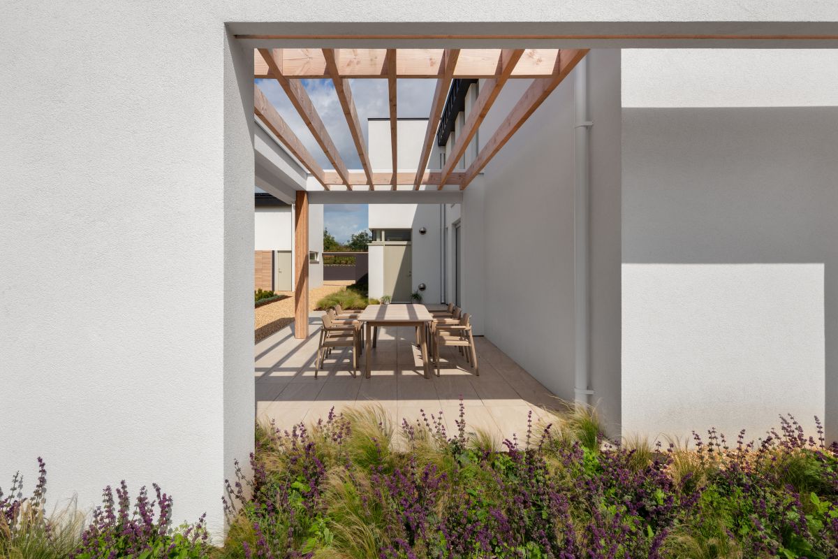 surrounded by elegant lightwood chairs. The lightwood flooring adds a touch of warmth to the space, complementing the natural surroundings. The white exterior walls, adorned with wooden framework overhead, create a charming setting. In the foreground, a plot of various grasses and plants adds a vibrant touch of greenery.