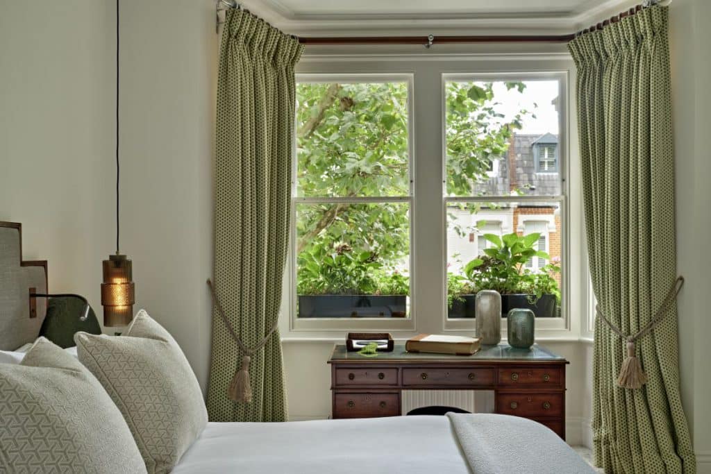 In this luxurious double bedroom with a beautifully styled bed. The pristine white bedsheets provide a clean and inviting canvas, complemented by the cream geometric Sophie Patterson scatter cushions that add a touch of elegance. A sophisticated grey fabric headboard with dark wood framing with built in book lights. An elegant throw elegantly falls ove the end of the bed .A captivating glass pendant light hangs either side of the bed . Natural light streams through the window, encasing the room in natural light. A dark wood desk sits in the window accompanied by a dark green velvet pouffe, complemented by a set of spotty green curtains.