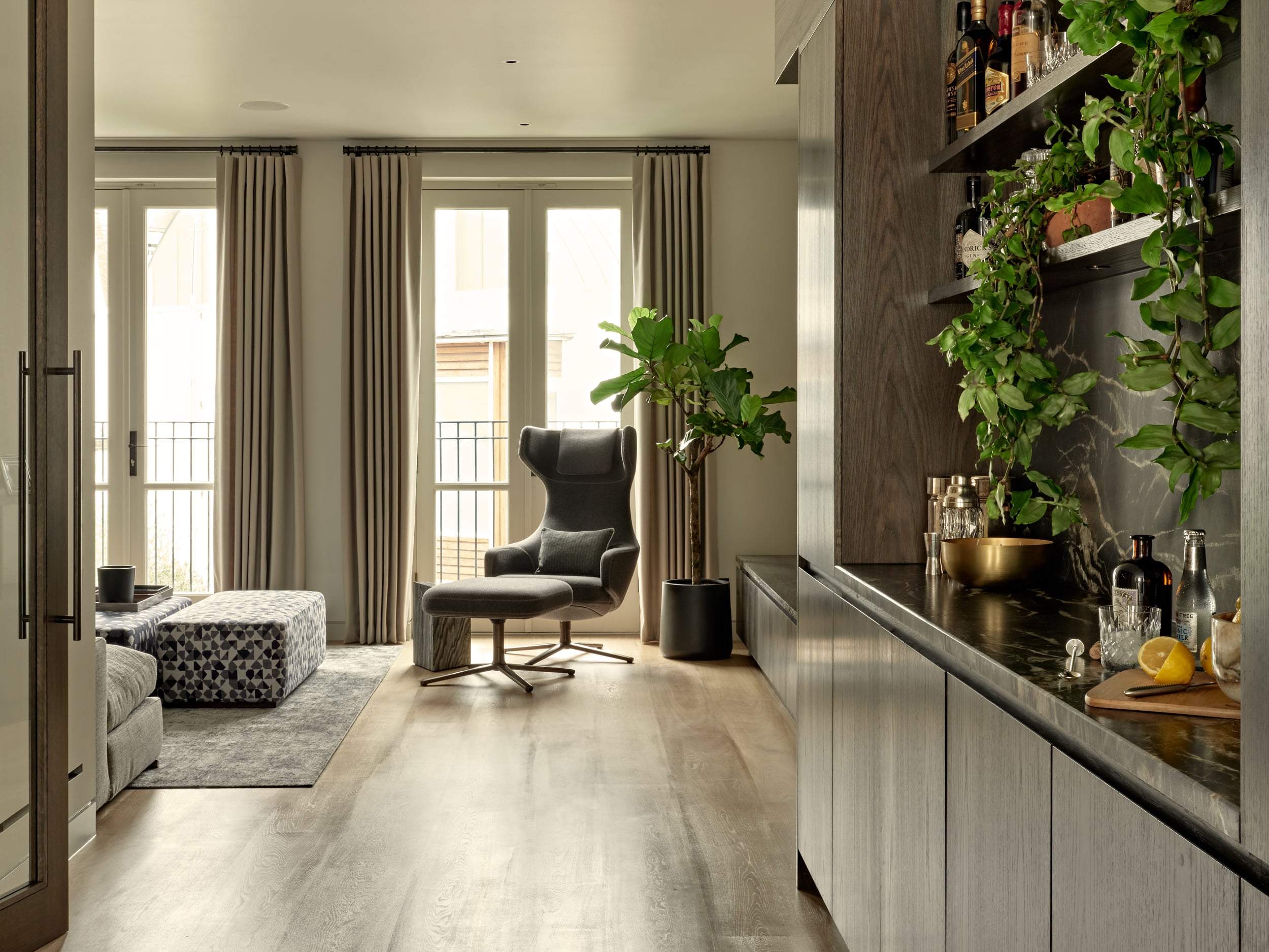 In a luxurious living room, the light wood flooring creates an inviting and warm ambiance. Large windows adorned with cream curtains bathe the space in natural light, enhancing the bright and airy atmosphere. The focal point is the Grand Repos armchair and ottoman by Vitra, upholstered in sleek grey fabric, offering a comfortable and stylish seating option. A wood block triangular side table stands adjacent to the chair, providing a convenient surface for placing items. A tall green plant adds a touch of freshness and natural beauty, while a wooden ottoman. On the opposite wall, a meticulously designed home bar with dark wood joinery offers a sophisticated and elegant spot for enjoying drinks and entertaining guests.