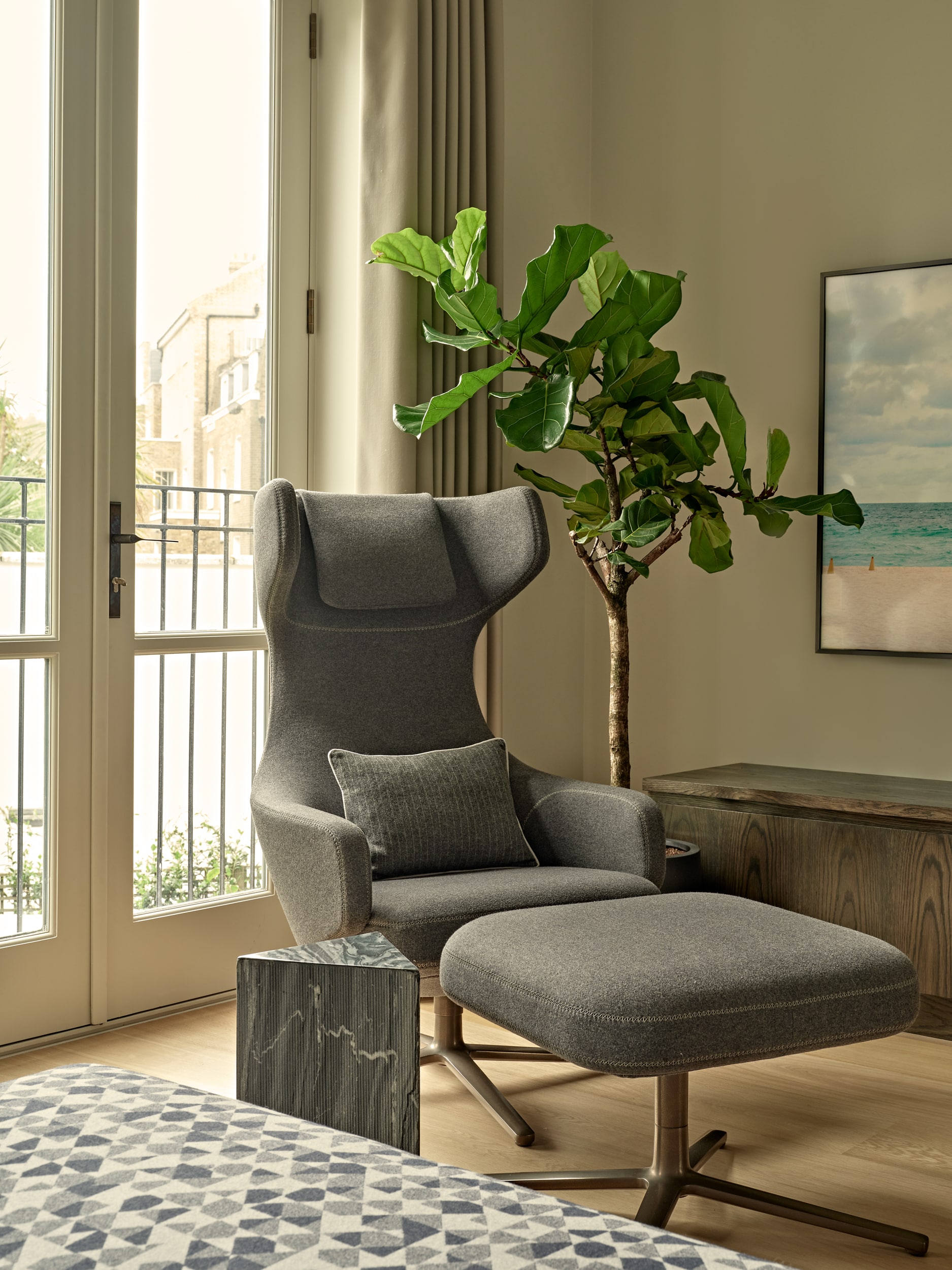 This luxurious media room/study exudes an elegant and serene atmosphere. The light wood flooring sets a warm foundation, complemented by large windows adorned with cream curtains that allow natural light to fill the space. The centrepiece is the Grand Repos armchair and ottoman by Vitra in a stylish grey hue, inviting relaxation and comfort. A wood block triangular side table stands alongside the chair, providing a convenient surface for books or beverages. A tall green plant adds a touch of nature, while a wooden ottoman with framed art above it completes the sophisticated ambience of this luxurious media room/study.