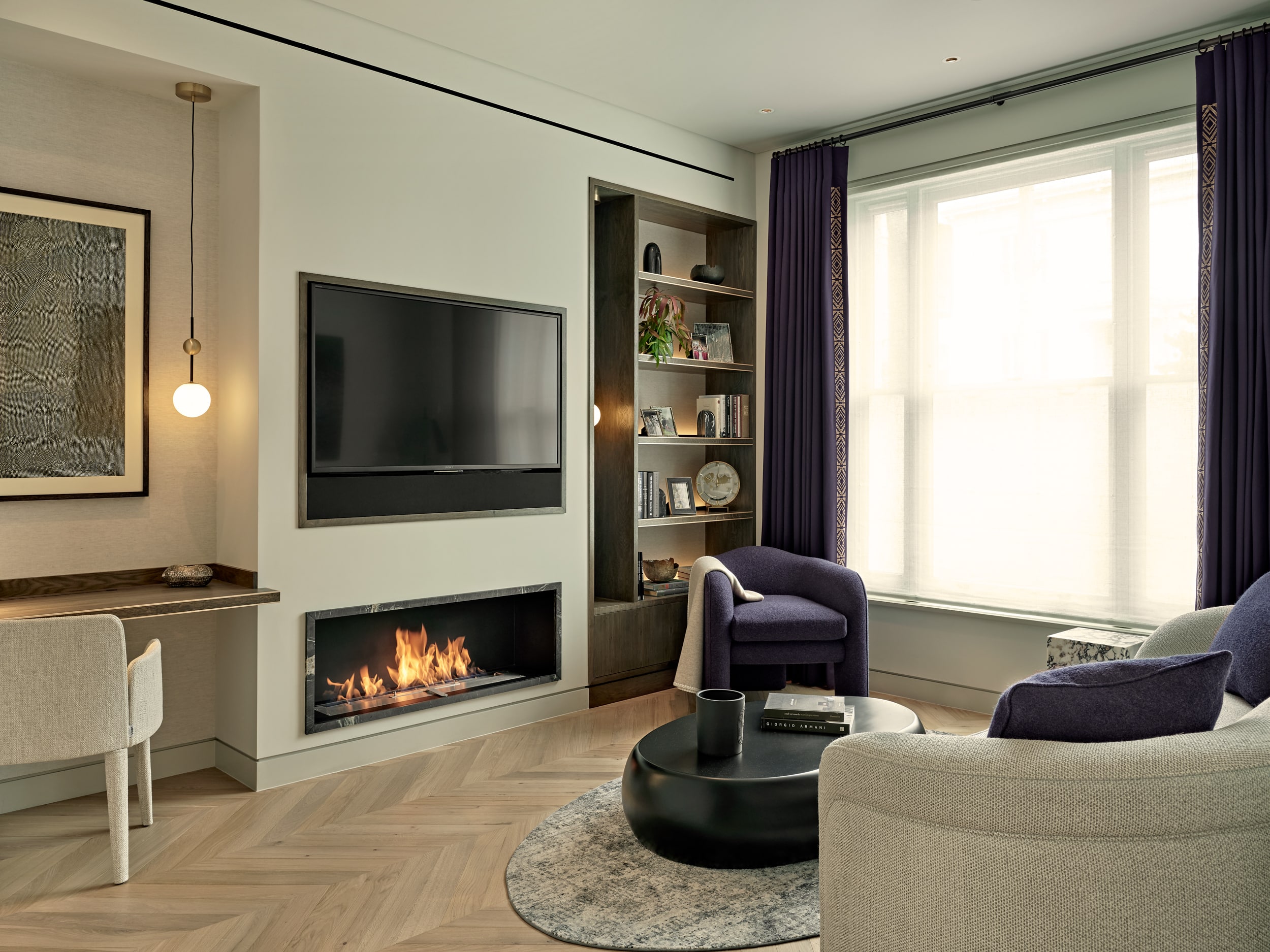 This modern luxury living room features lightwood herringbone flooring. A wall-mounted TV sits above a sleek black marble-framed fireplace. On the right-hand side of the TV, dark wood shelving is built into the wall, adorned with books. In the centre of the room, a cosy curved sofa with purple accent cushions is complemented by a matching armchair. They rest on a white and grey circular rug with a simple pattern, accompanied by a black circular coffee table displaying books. Adjacent to a large window dressed in white blinds and purple curtains, a built-in dark wood desk space with a cream textured chair and a hanging sphere light provides a stylish workspace. Art pieces are thoughtfully mounted on the walls.