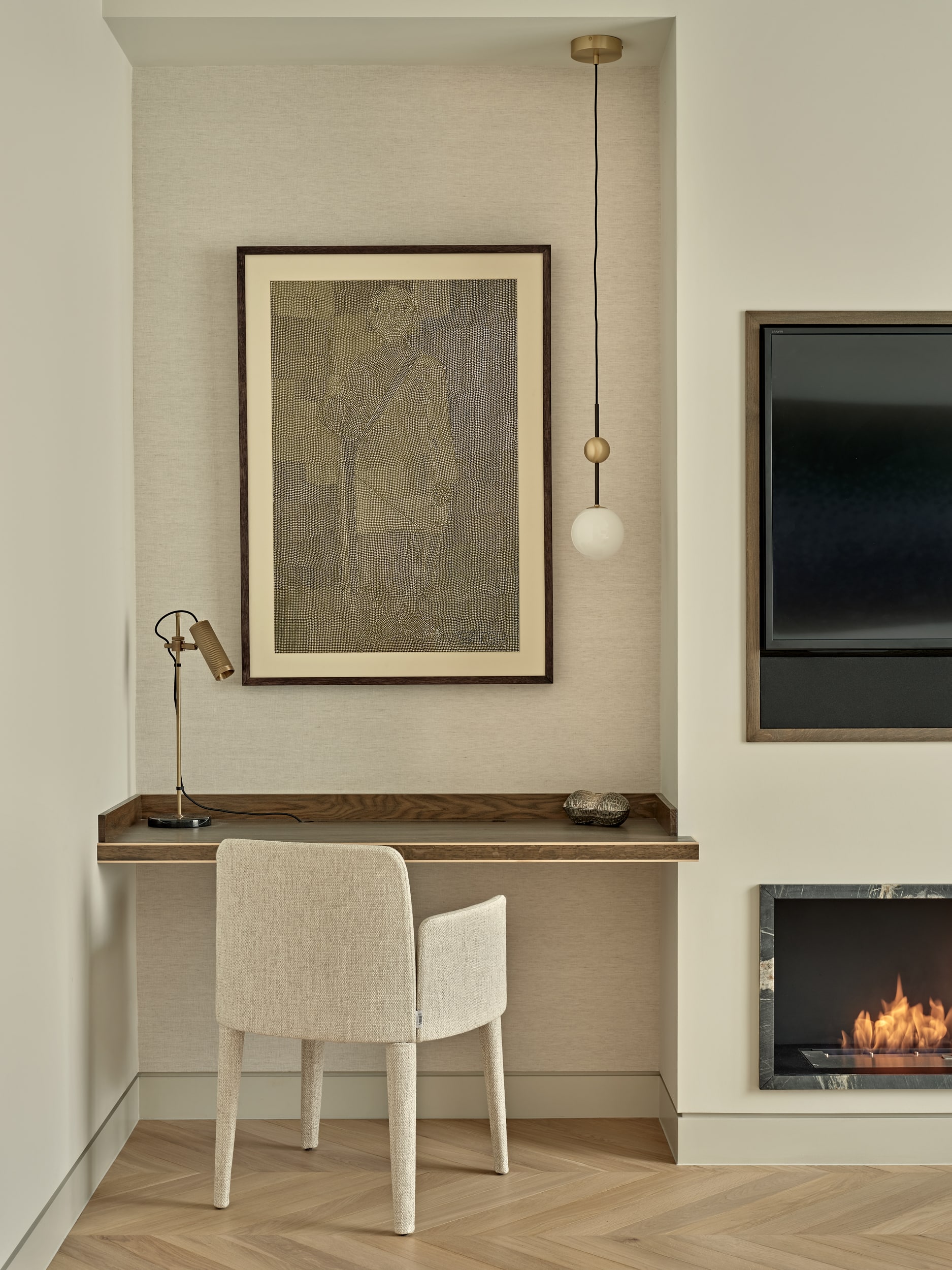 luxury, thoughtfully designed study area, nestled in the corner of this living room. With fresh white walls and a section of textured cream wallpaper behind the desk. The dark wooden desk, seamlessly built into the nook, provides a convenient workspace. A comfortable cream textured chair tucks neatly underneath the desk. A gold table lamp adds a touch of style to the desk surface. Above the desk, an art piece by renowned Indian artist Sheshdhar Pandeycreates a focal point. Adding elegance to this space, a duo of cylinder hanging lights one as a light function, the other adorned with a golden cylinder for decoration, balancing functionality and aesthetics. Adjacent to the desk area, a mounted TV above a built in fireplace linking you to the living area.