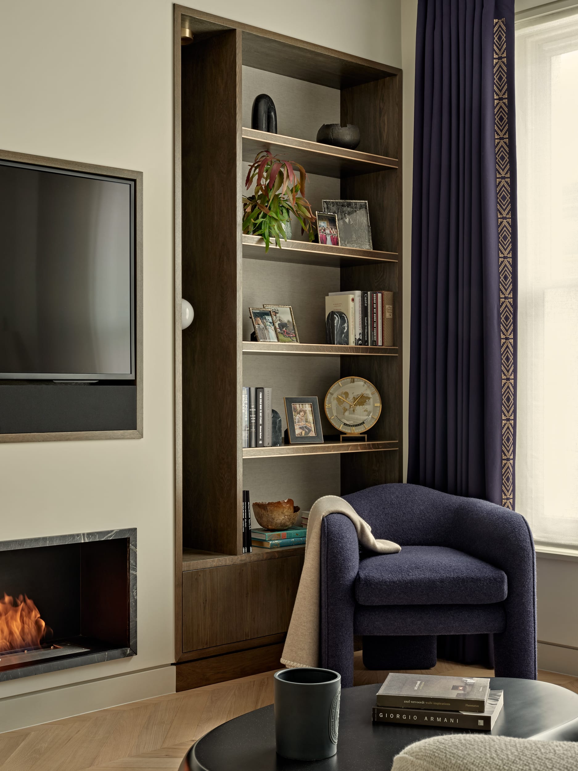 This modern luxury living room features lightwood herringbone flooring. A wall-mounted TV sits above a sleek black marble-framed fireplace. On the right-hand side of the TV, dark wood shelving, by Lethbridge bespoke joinery, built into the wall, adorned with books. In the centre of the room, Ben Whistler bespoke, purple armchair with a cream throw draped over one arm, accompanied by a black circular coffee table displaying books. Adjacent to a large window dressed in white blinds and purple curtains.