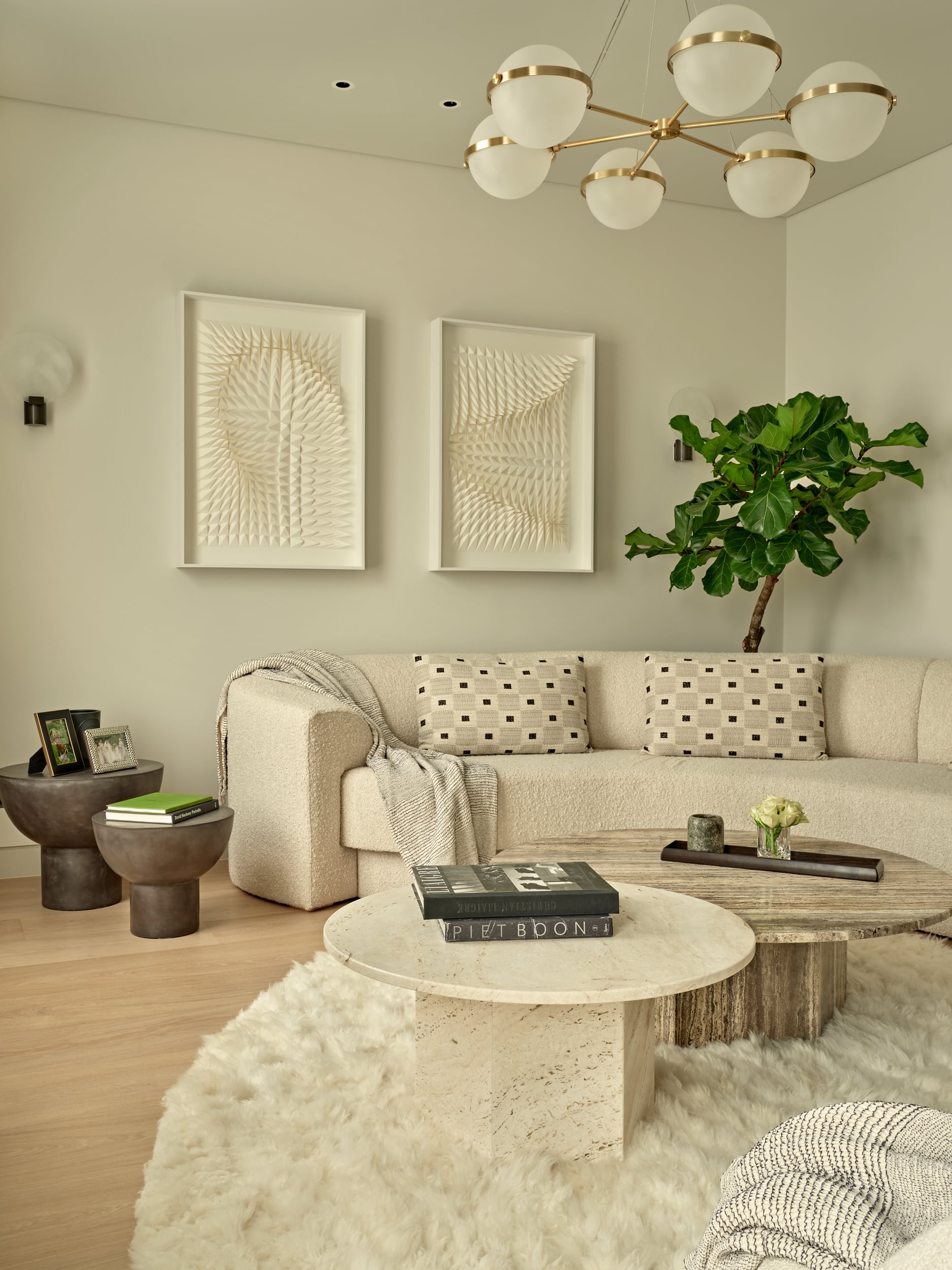 This luxurious living room features a corner space adorned with a curved white, textured sofa. Three matching white cushions and a white throw add comfort and elegance. A fluffy curved rug lies in front of the sofa, hosting two marble tables in different natural tones. Adjacent to the sofa, two black side tables display books and framed photos. Adorning the wall are two white textured art pieces, while behind the sofa stands a tall, vibrant green plant, adding a touch of nature to the space.