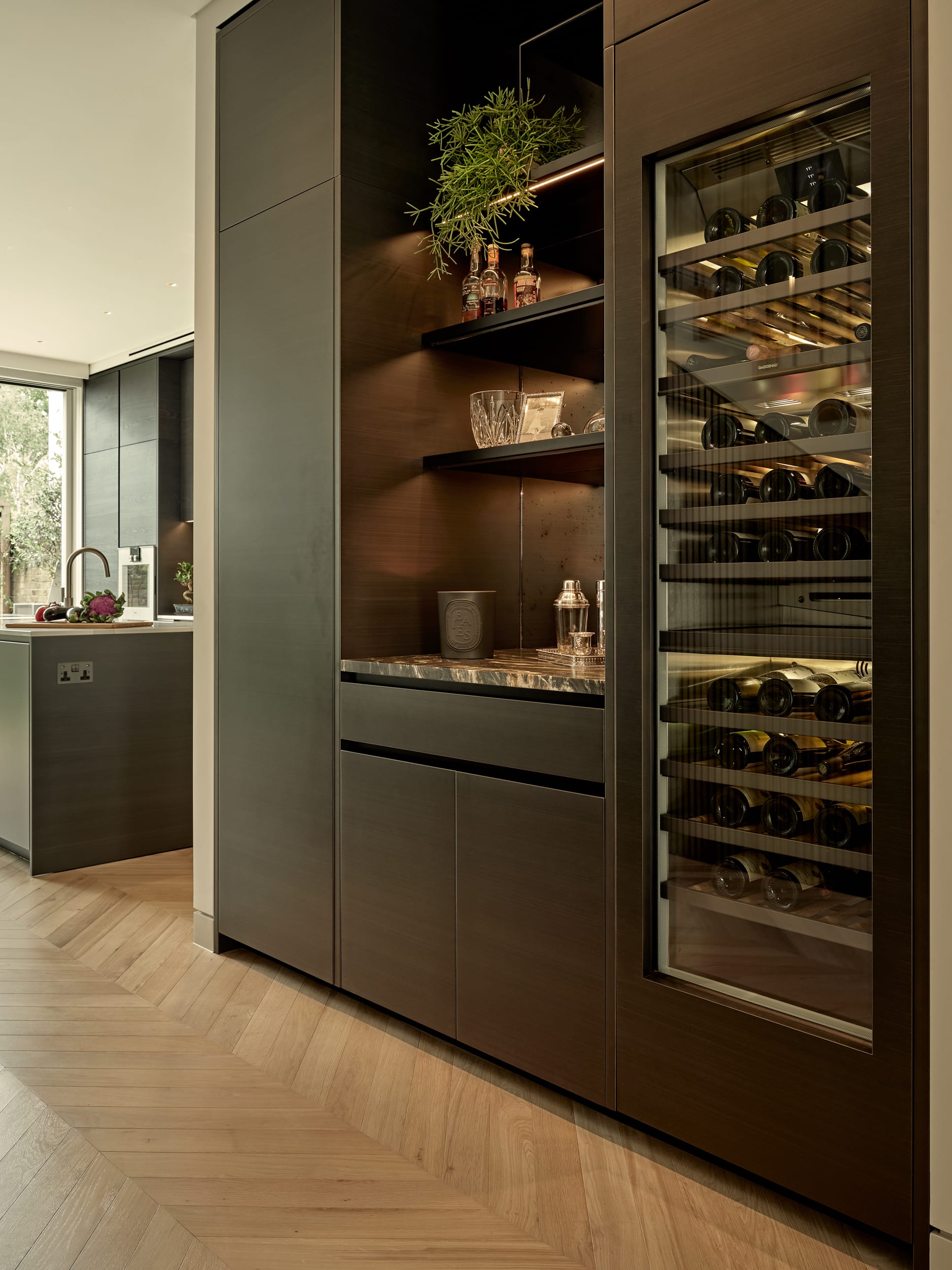 A luxurious home bar with its light wood herringbone floor. The bar exudes elegance with its sophisticated design and attention to detail. A wine fridge, stocked with an impressive selection of bottles. The dark wood joinery, complemented by a pristine marble countertop, creates a striking focal point. Adorned with cocktail shakers and a wine ice bucket. Above the bar, shelves showcase an assortment of small liquor bottles, glassware, and green plants, adding a touch of natural beauty. beyond the bar, the space transitions into the inviting kitchen.