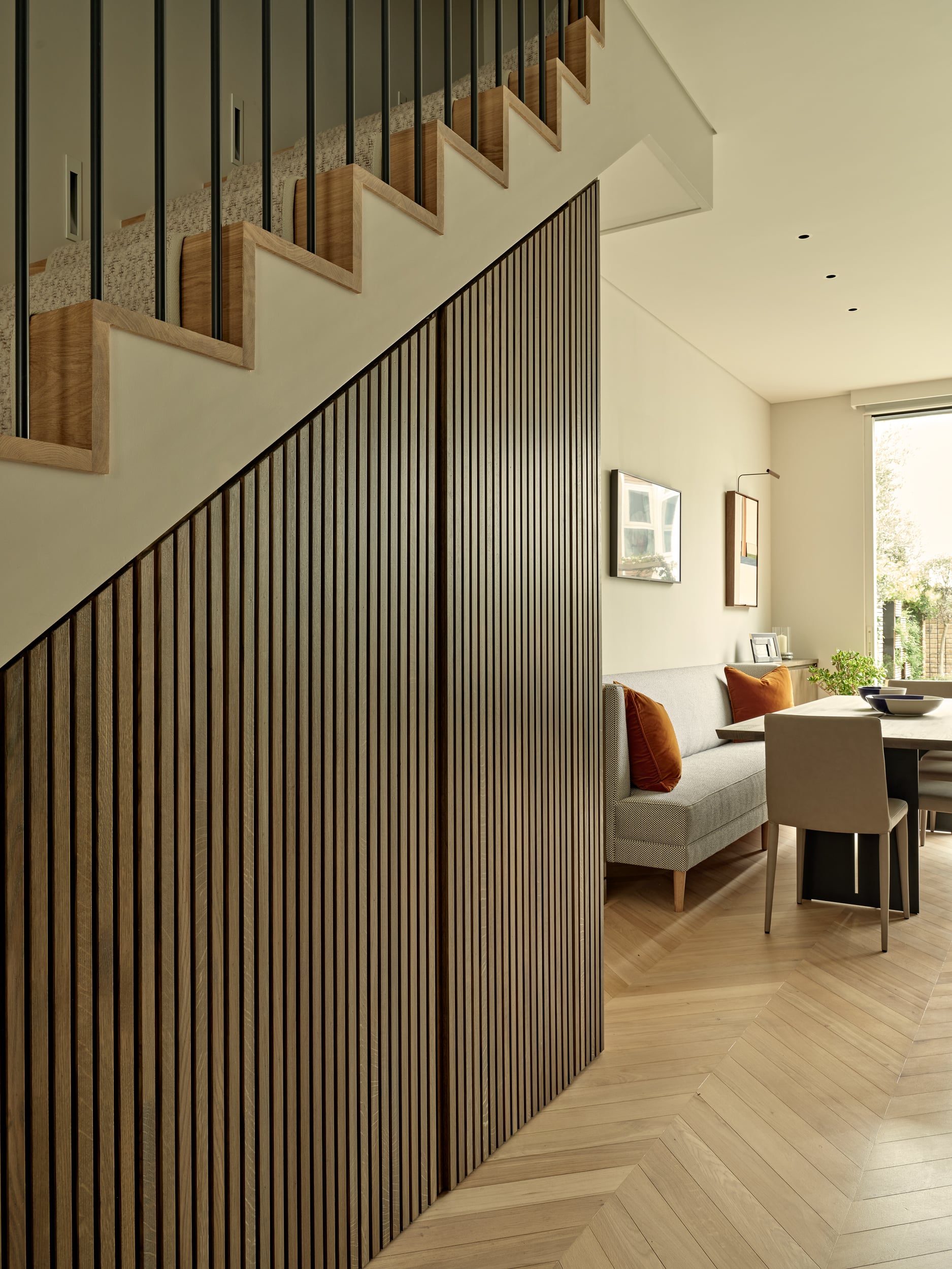 This zoomed-in image unravels the beauty of a luxury stairway. Thin wood panelling graces the wall beneath the stairs, concealing hidden storage that enhances both functionality and aesthetics. The stairs themselves are crafted from light wood and are adorned with beige carpet. Beyond the stairs lies an inviting dining space, featuring light wood herringbone flooring and pristine white walls and ceiling. A sophisticated grey dining couch with velvet orange cushions, while a light wood dining table adorned with tasteful bowls takes centre stage. A white chair stands at the end of the table. Above the couch, two captivating pieces of art captivate the senses, infusing the space with artistic allure.