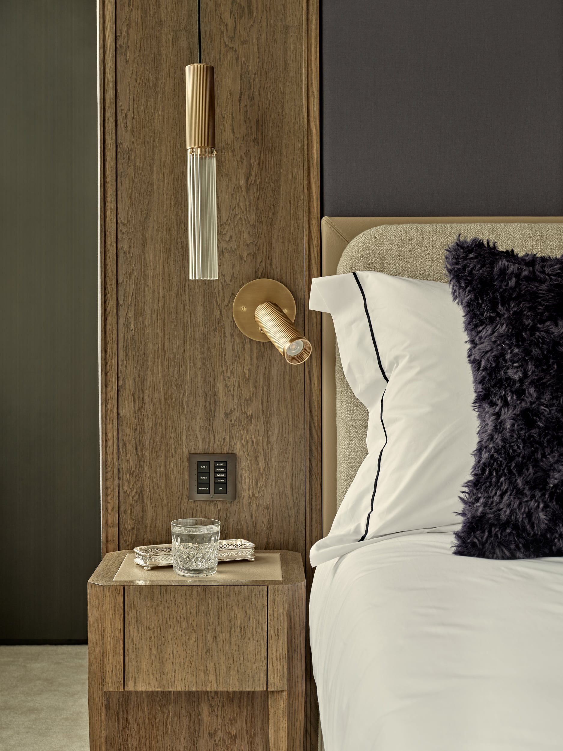 This high-end bedroom showcases a luxurious ambiance with its carefully curated design elements. A dark wood room divider serves as an elegant backdrop to the double bed, while matching bedside tables add a cohesive touch. The grey/blue fabric wall panelling behind the bed's headboard creates a soothing focal point. A natural cushion headboard enhances the comfort and style of the double bed, complemented by white cotton bedsheets with blue/grey detail edging. Fluffy blue/grey accent cushions add a touch of plushness, while hanging lights on either side of the bed provide atmospheric illumination alongside wall-mounted book lights.