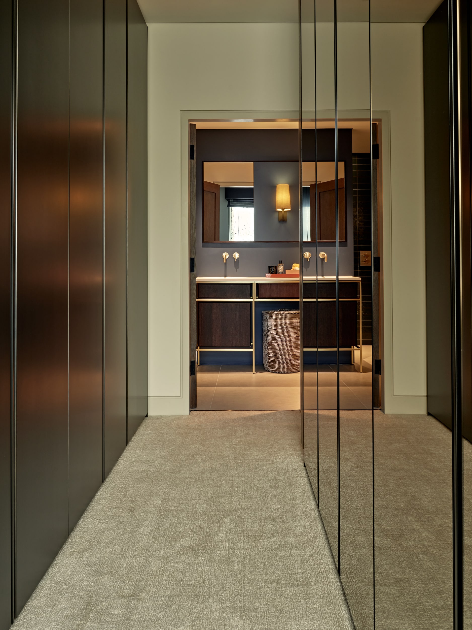 This dressing room leading to an exquisite ensuite. The room exudes sophistication with its contrasting elements. On one side, sleek matt black wardrobe doors create a stylish storage solution, while on the opposite wall, mirror vanity wardrobe mirrors add elegance and functionality. At the end of the room, a glimpse into the ensuite reveals a captivating scene. The ensuite boasts a blend of dark wood and gold accents, with a stunning sink area adorned with gold taps and a square mirror above. Soft blue-grey walls complete the serene and opulent ambiance of this enchanting space.