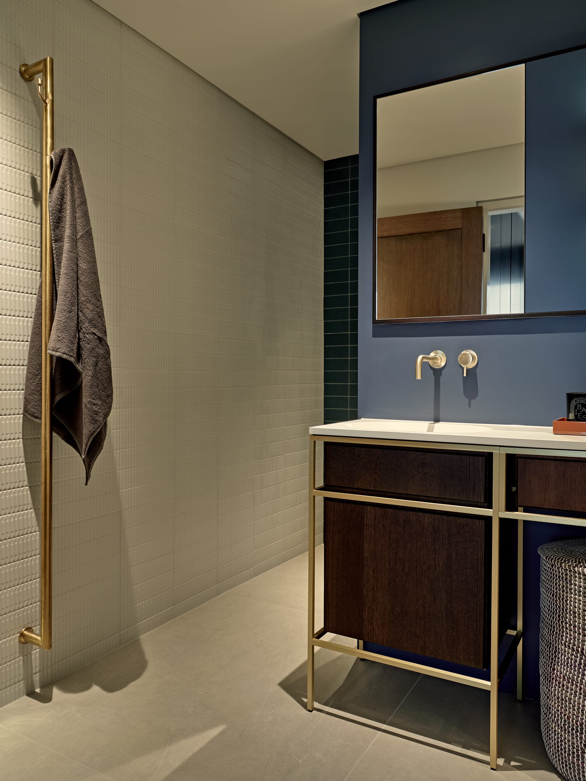 This is the epitome of a luxury bathroom. The grey stone tiled floor sets the stage for a serene atmosphere, complemented by the crisp white textured Marca Corona Regoli Porcelain wall tiles. A touch of elegance is added with the dark teal wall tiles on one side, while the sky blue painted wall adds a pop of bright colour. A centrepiece in the room, a large rectangular mirror hangs above a white sink with a brown wood base and gold metal pole structure, harmonising with the gold accented taps and towel rail. A dark grey towel hangs from the towel rail adding a homely, functional aspect to the setting.