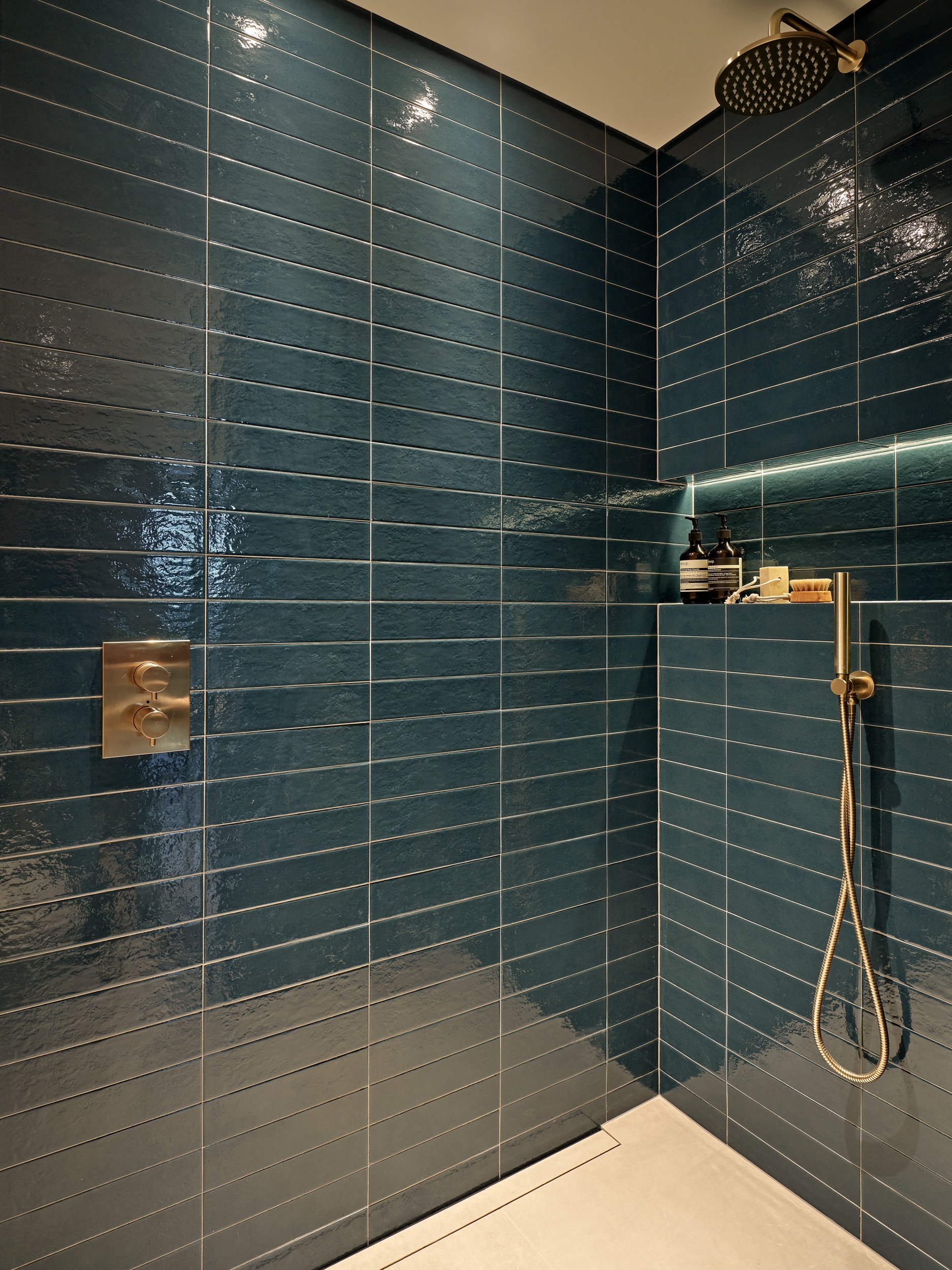 up-close image of a shower. The shower showcases deep blue subway tiles from the prestigious Collection Refoli by Marca Corona, exuding elegance and style. The gold brushed brass hardware, including the shower head and control panel, adds a touch of sophistication. Complementing the shower, light cream floor tiles create a striking contrast. A built in shelf halfway up the wall with built in lights, illuminating the shelf and displaying an array of shower products.