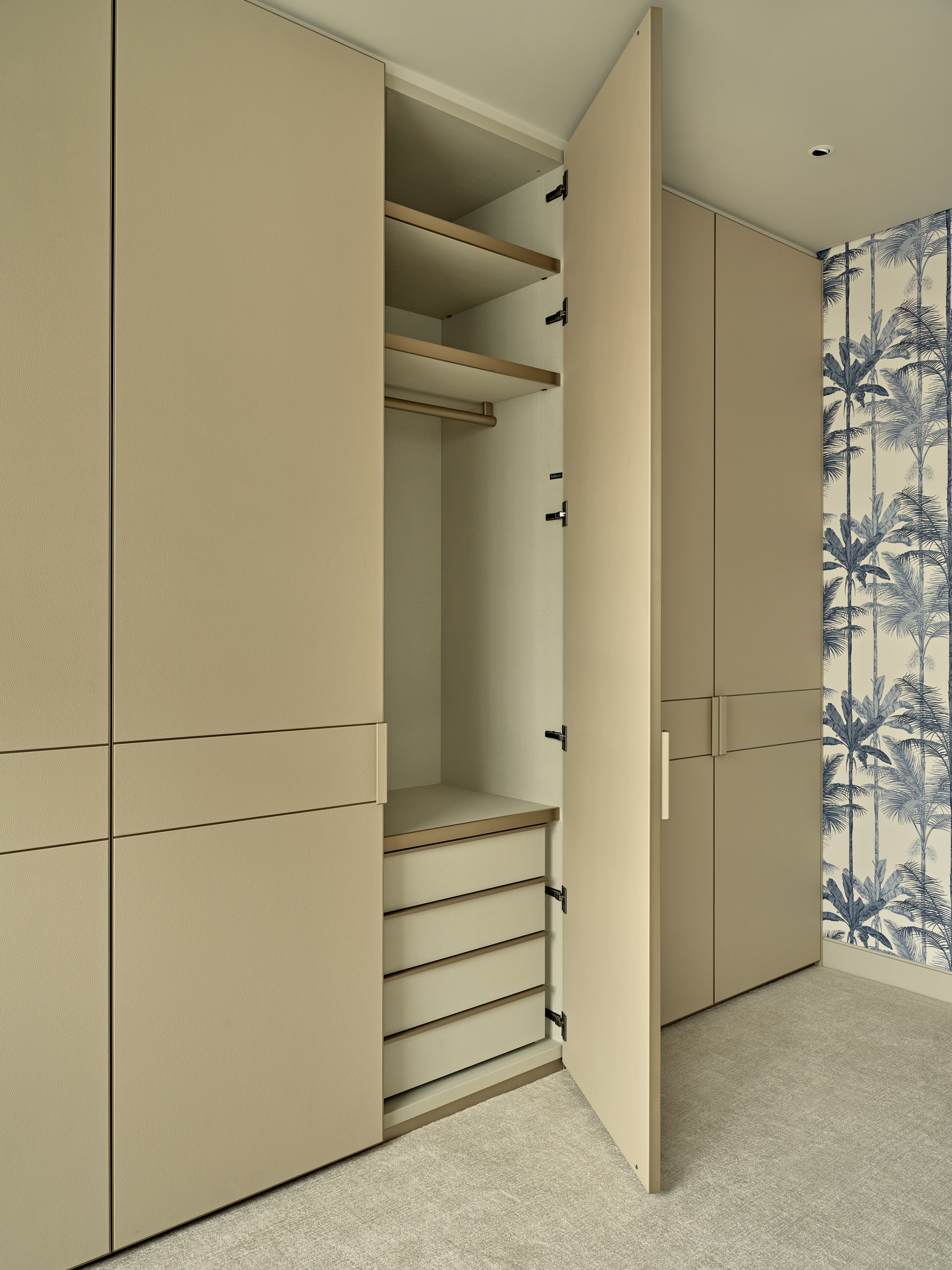 This luxurious bedroom/dressing room offers a harmonious blend of elegance and functionality. The wardrobe joinery is adorned in a soothing beige leather, with one open door revealing a well-organised selection of storage options. A plush beige woven carpet enhances the comfort and style of the space. The walls are adorned with a striking blue and white wallpaper featuring palm trees and plants, creating a captivating tropical ambiance. This combination of tasteful design elements transforms the room into a serene and sophisticated retreat.