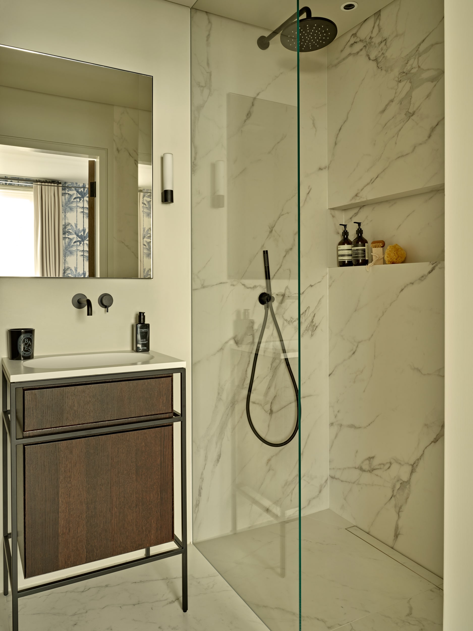 Luxury bathroom adorned with white marble featuring grey veins. The shower has a sleek glass screen and black hardware, complemented by a built-in shelf adorned with bathroom accessories. To the left, a white sink with a dark wood and black metal base, accompanied by a wall-mounted mirror and subtle wall lights on either side.