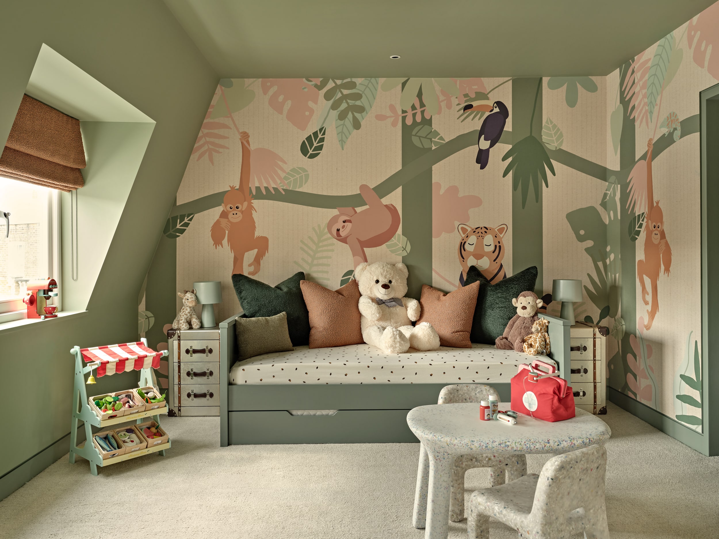 Welcome to the luxury kids' playroom, transformed into a vibrant jungle paradise. A green trundle bed adorned with burnt orange and green cushions is accompanied by stuffed jungle animals. The walls are adorned with wallpaper showcasing lush jungle scenery and playful animals. The green-painted walls create a lively atmosphere. Burnt orange blinds adorn the window, adding a pop of colour. Green old suitcase-style side tables with matching lamps provide storage and illumination. A toy food shop and toy coffee machine offer endless opportunities for creative play. A children's drawing table with two seats inspires artistic exploration. This jungle-themed playroom is a haven of fun and creativity for children to explore and let their imaginations roam.