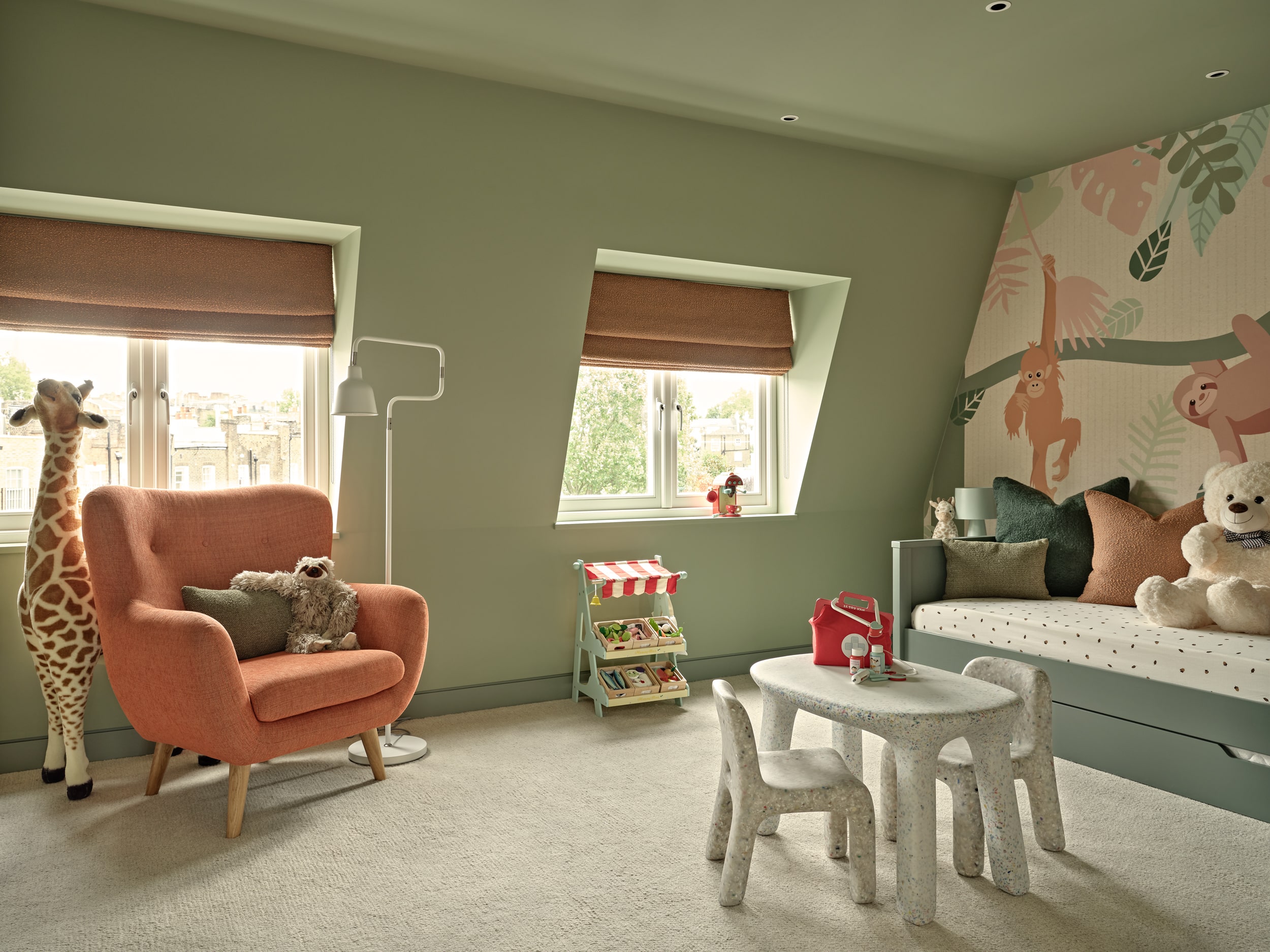 A luxury kids' playroom, transformed into a vibrant jungle paradise. A green trundle bed adorned with burnt orange and green cushions is accompanied by stuffed jungle animals. The walls are adorned with wallpaper showcasing lush jungle scenery and playful animals. The green-painted walls create a lively atmosphere. Burnt orange blinds adorn the window, adding a pop of colour. Green old suitcase-style side tables with matching lamps provide storage and illumination. A toy food shop and toy coffee machine offer endless opportunities for creative play. A children's drawing table with two seats inspires artistic exploration. This jungle-themed playroom is a haven of fun and creativity for children to explore and let their imaginations roam. Adjacent to this, a cosy orange armchair accompanied by a small green accent cushion and a toy sloth, with a white floor lamp for reading carefully positioned . Behind the armchair a large toy giraffe stands tall bringing this cosy corner to life.