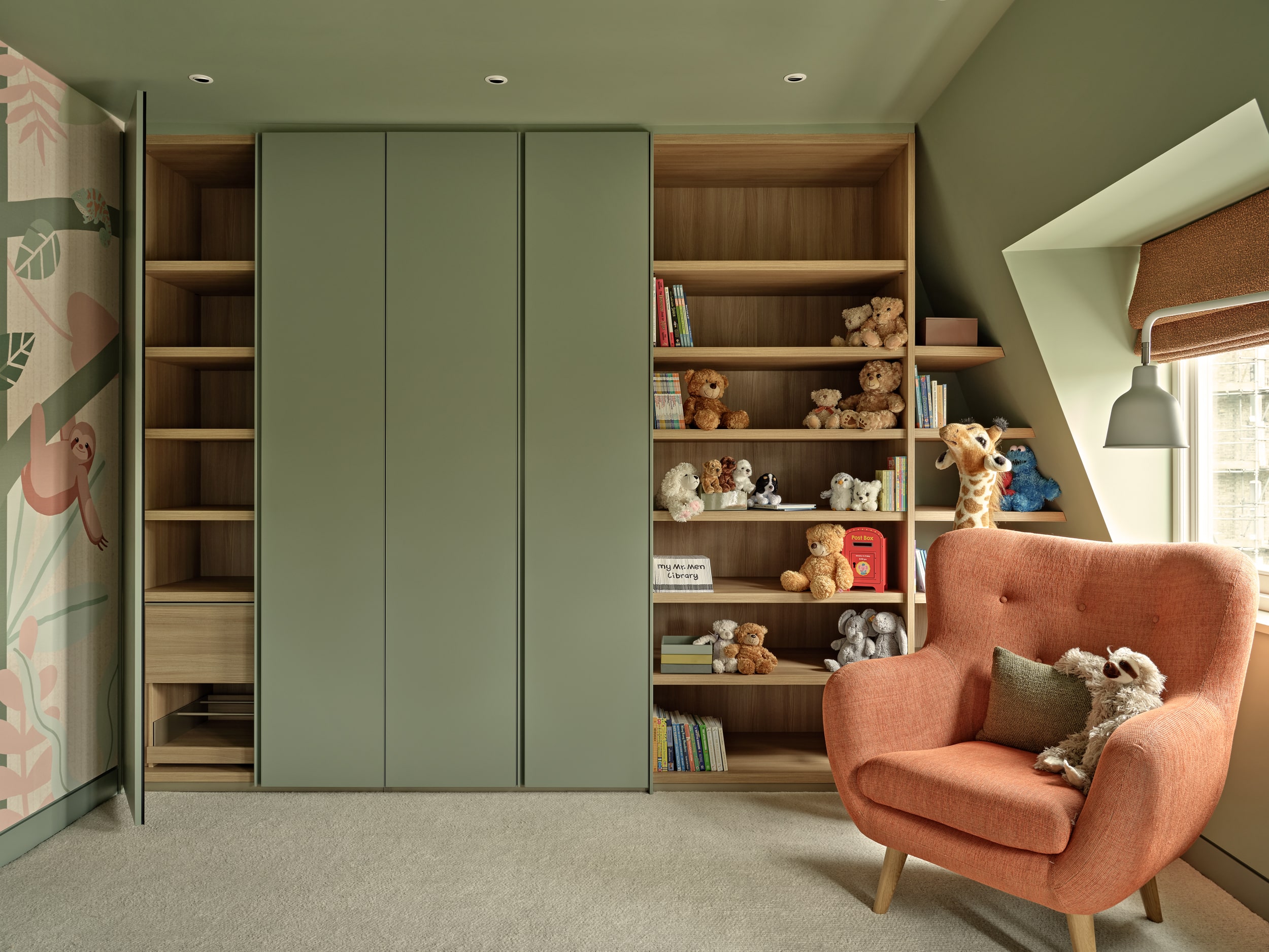 A luxurious children's playroom where creativity and comfort come together. The cream carpet sets the foundation for a cosy atmosphere, while the green walls add a touch of vibrancy. Along the back wall, bespoke joinery provides wardrobe space, alongside shelves that display a collection of colourful books and stuffed toys. A window adorned with textured orange blinds invites in natural light. In front of the window, a plush orange armchair with a green accent cushion becomes the perfect spot for reading, accompanied by a charming toy sloth. Standing tall behind the armchair, a playful toy giraffe adds a whimsical asset.