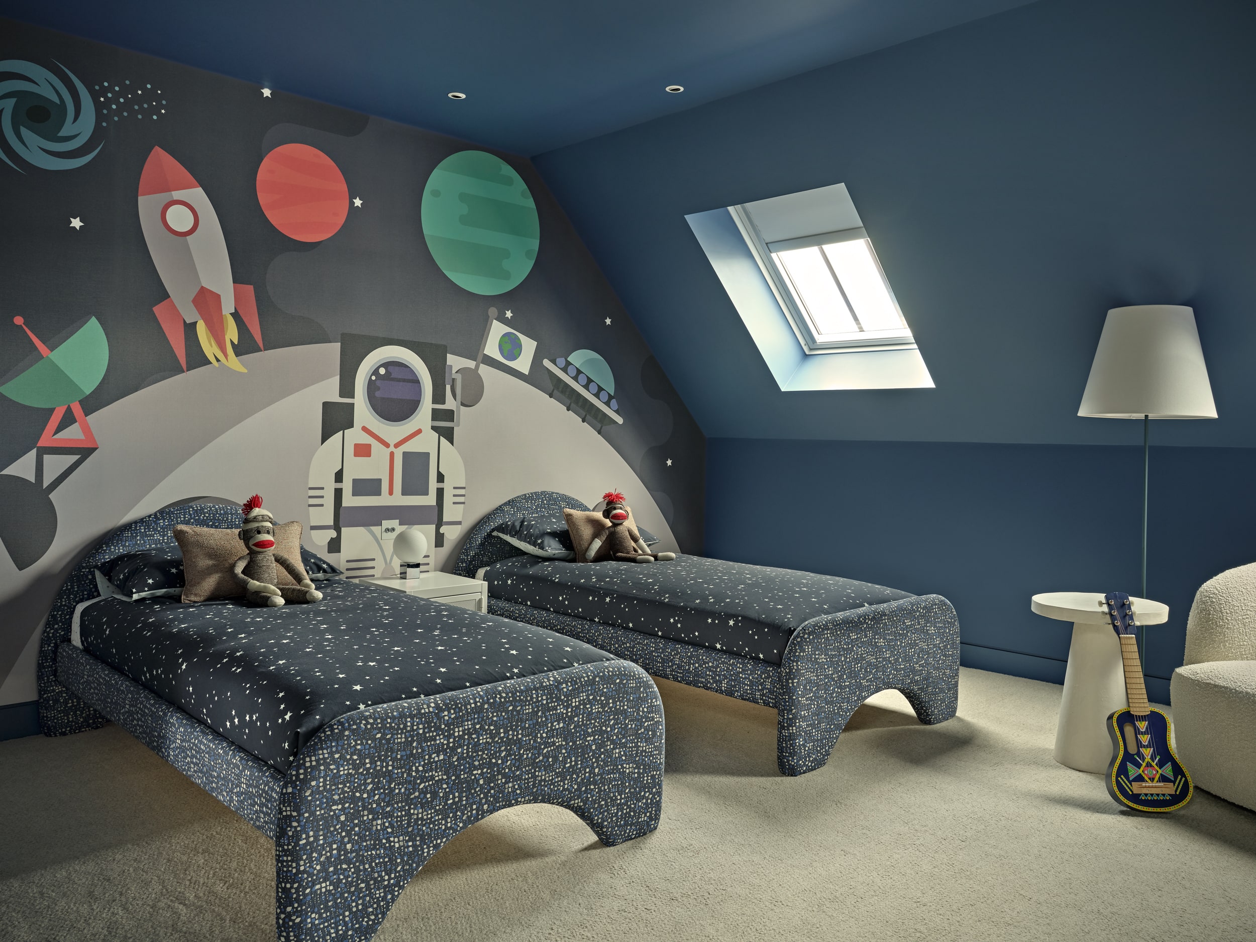 A twin children's bedroom, a captivating space-themed wall mural sets the stage for cosmic adventures. Two single twin beds beckon with their charming blue patterned fabric headboards and bed bases, adorned with blue and white starry bedding. Toy monkeys accompany each bed, adding a touch of personality. Blue-painted walls and ceiling, enhanced by a skylight that invites natural light to dance across the room. A cosy white fluffy armchair nestled in the corner invites you to curl up with a book, while a white floor lamp and side table create a cosy reading nook. A small blue guitar leans against the side table.