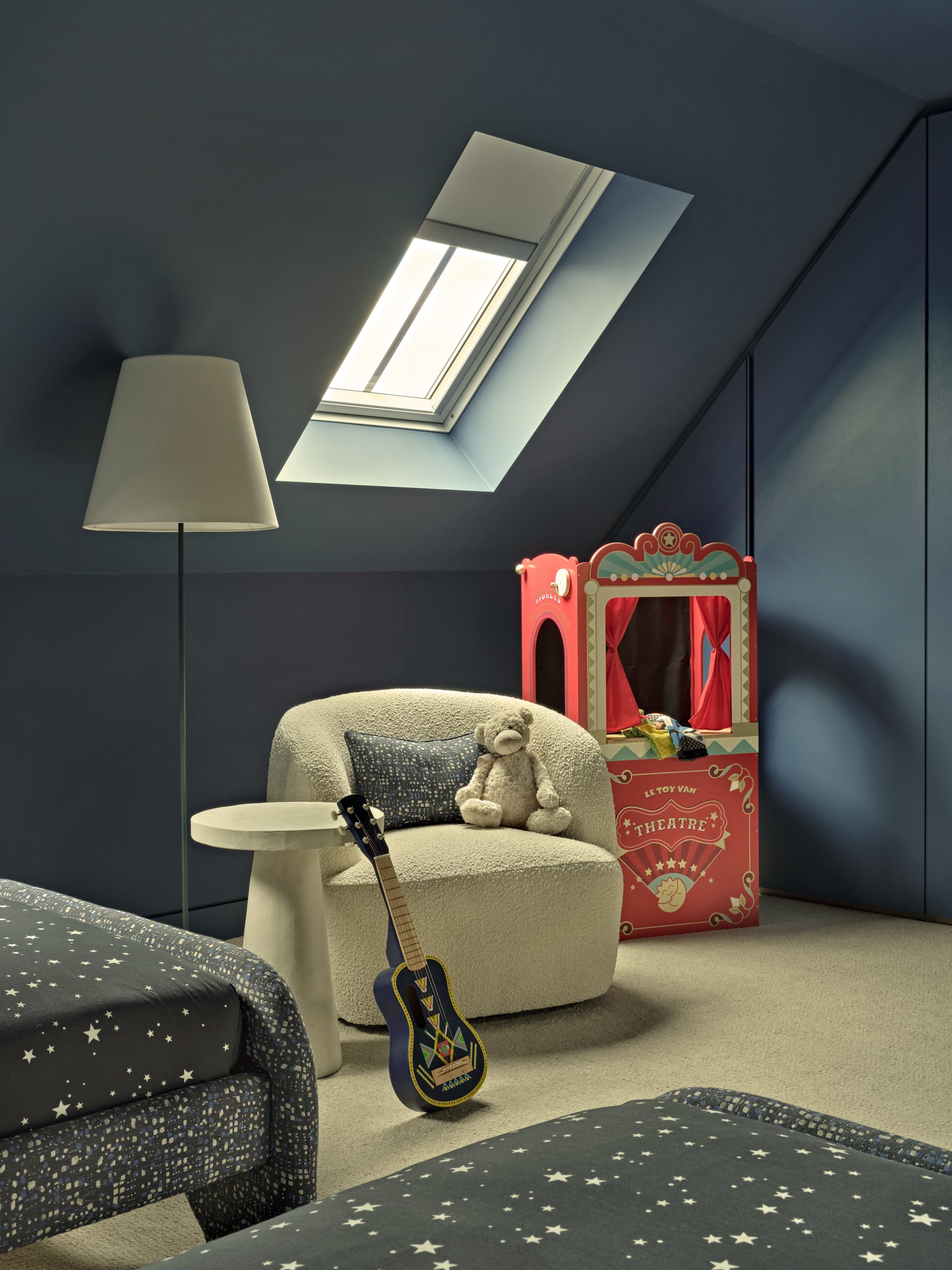 This luxurious kids' bedroom/playroom features calming blue painted walls illuminated by a skylight. A plush cream carpet adds comfort and warmth to the space. Two blue single beds adorned with bedding featuring white stars create a dreamy sleeping area. A cream fluffy, gwyneth boucle armchair with a matching blue cushion complements the fabric on the bed bases. A white tall side table holds a small blue guitar, while a white floor lamp provides additional lighting. In the corner of the room, a vibrant red mini puppet theatre awaits imaginative play and storytelling adventures.