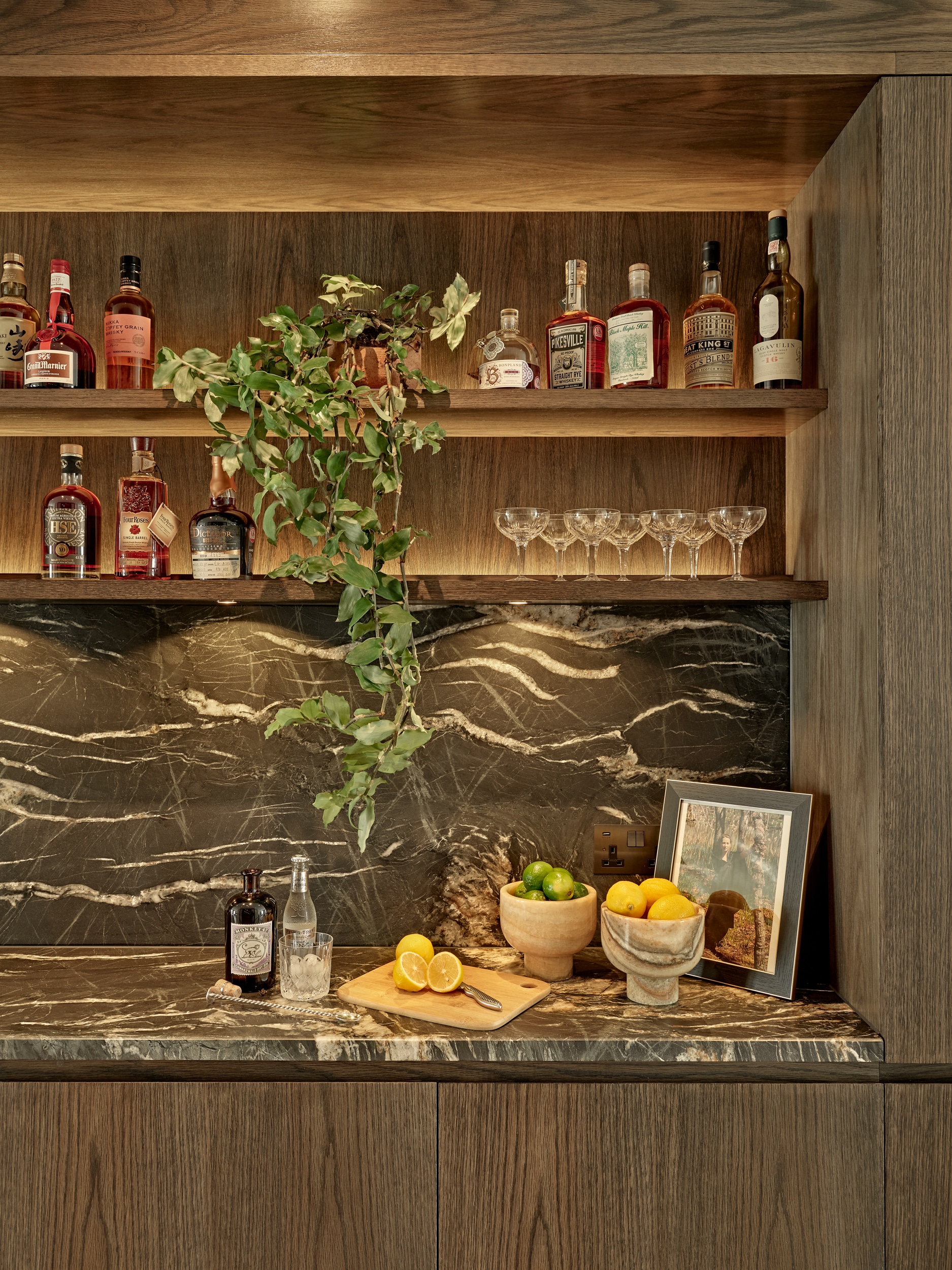 A luxurious modern home bar, designed for cocktail enthusiasts. The dark wood joinery features illuminated shelving, showcasing an array of meticulously displayed liquor bottles and glasses. A stunning grey marble countertop with elegant white veins adds a touch of sophistication. On the countertop, a selection of fresh lemons and limes are accompanied by a cutting board, ready for the creation of refreshing drinks. This luxury home bar seamlessly combines functionality and style, providing a sophisticated space for mixing and enjoying cocktails.