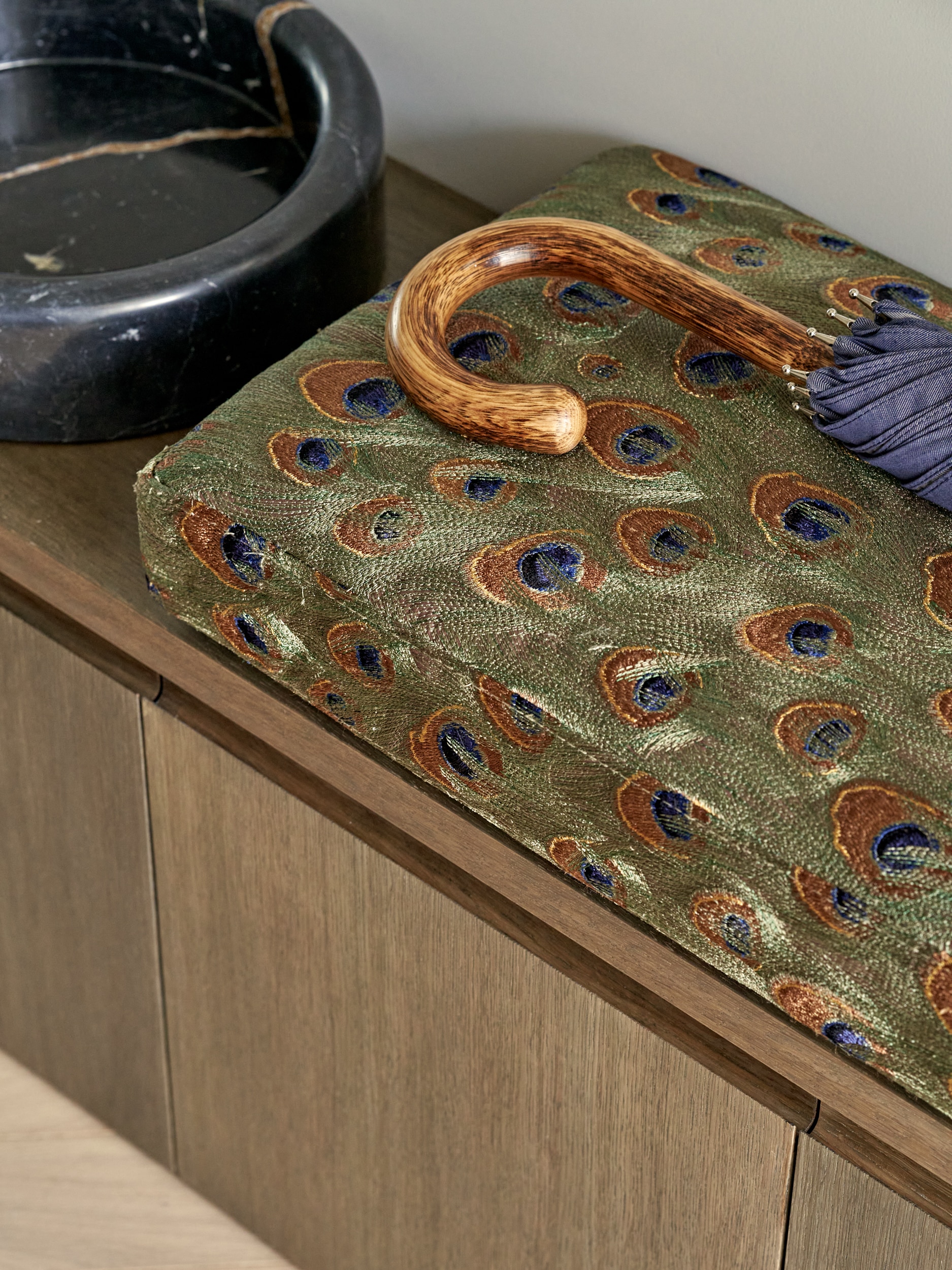 An up-close image of a luxurious area. A dark wood bench with drawers exudes functionality. Adorned with a plush cushion made out of a stunning peacock feather pattern. Alongside the cushion rests a wooden-handled umbrella. Adding a touch of sophistication, a black marble pet bowl sits nearby.