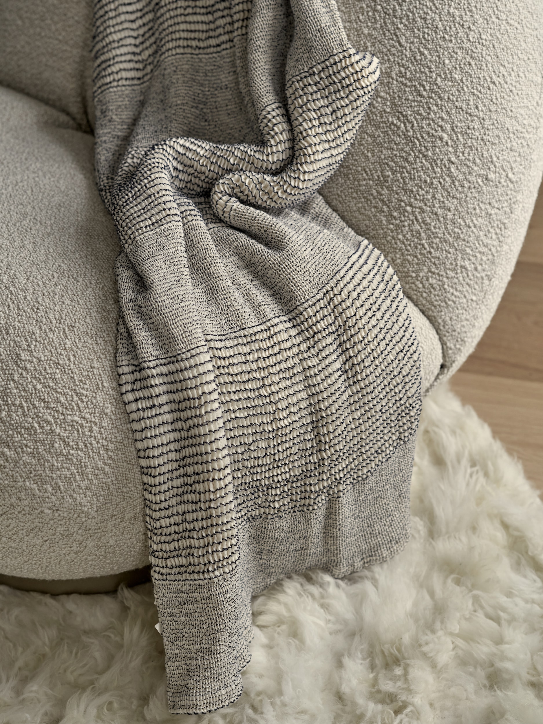 This luxurious living room captures a cozy and elegant atmosphere. A zoomed-in picture reveals a white Pacha Armchair adorned with boucle fabric, placed on a plush white fluffy rug. The light wood flooring adds warmth and sophistication to the space. A stylish throw in shades of grey and white enhances the comfort of the armchair.