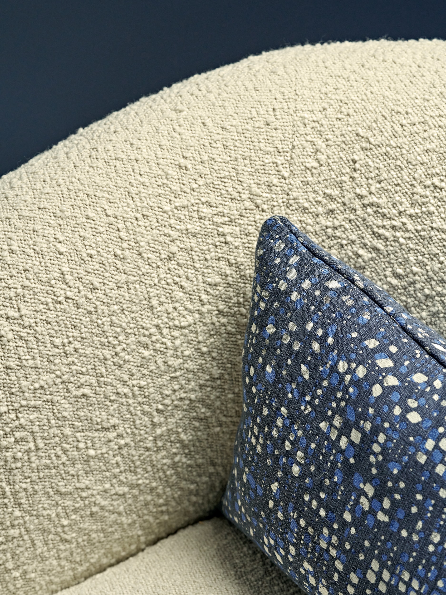 Indulge in the luxury of a zoomed-in image showcasing an armchair adorned with exquisite cream boucle fabric from CB2 America. The attention to detail is evident in the accent cushion, crafted from a dark blue fabric adorned with intricate patterns of light blue and beige. The combination of textures and colours creates a captivating and sophisticated aesthetic.